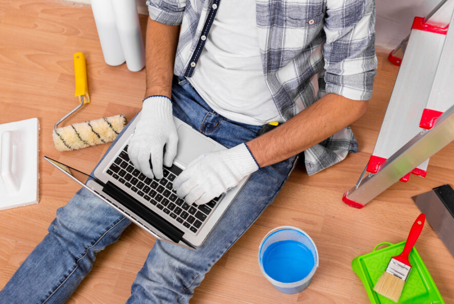 A handyman in Chicago sitting on the floor surrounded by painting tools, wearing gloves and casual work clothes, working on a laptop to plan or manage his tasks.