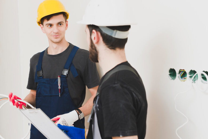 Two electricians wearing safety helmets discussing work near exposed wiring in a wall.